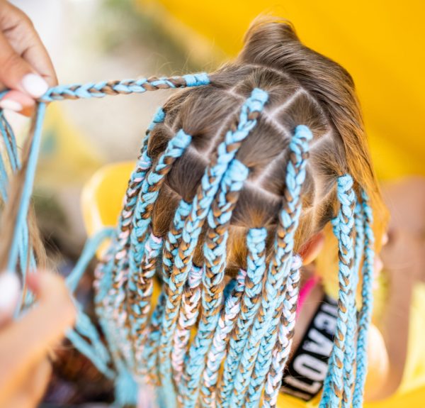 A little waiting tanned girl in a bright yellow summer suit, weaves colored pink-blue African braids in her dark wet thick hair on a hot sunny sea day