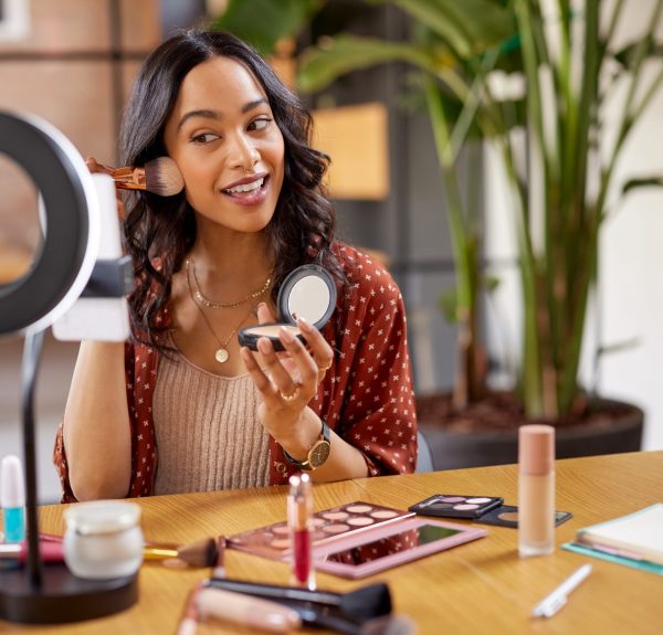 Young woman applying makeup on face with brush while recording vlog on smartphone for social media with LED ring lamp on table. Multiethnic fashion blogger creating cosmetic video. Professional hispanic beauty makeup artist vlogger livestreaming makeup tutorial while reviews a new blush product.