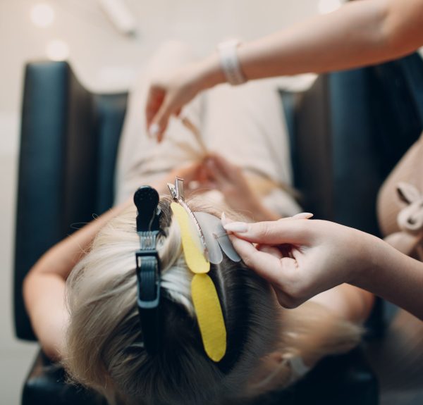 Hairdresser female making hair extensions to young woman with blonde hair in beauty salon. Professional hair extension