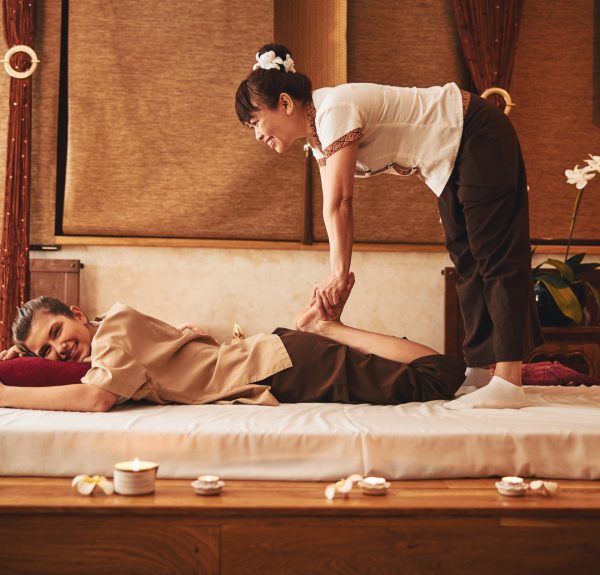 Female standing with legs on floor mattress and bending over lying woman while kneading her feet during Thai massage
